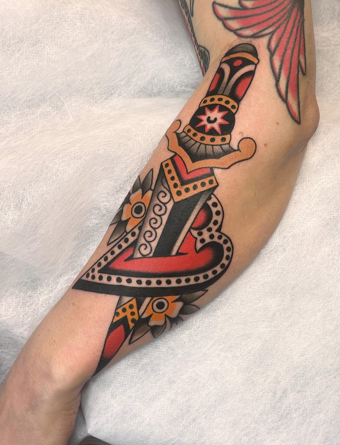 Colorful dagger tattoo on an arm featuring red, orange, and black details with decorative patterns and floral elements.