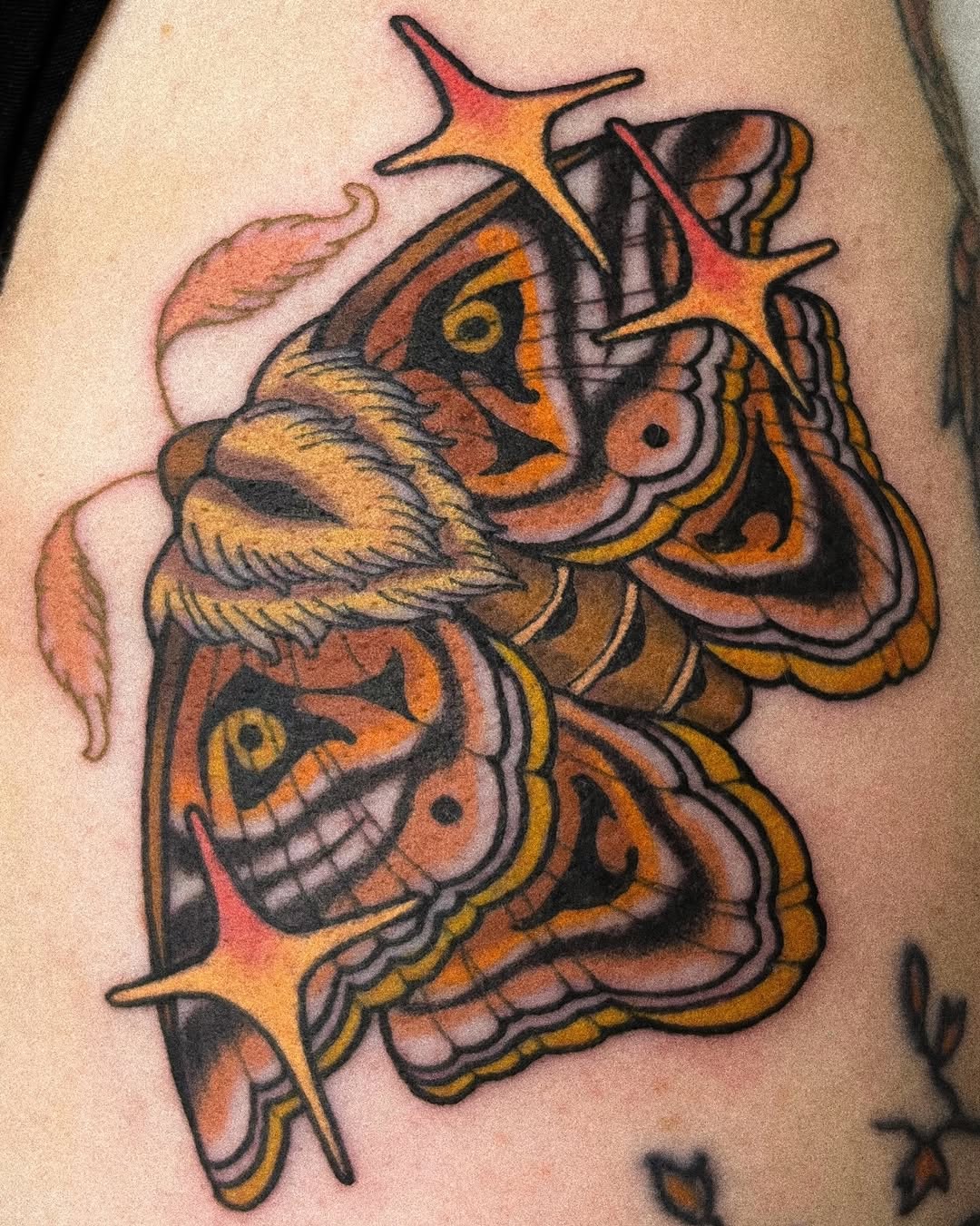 Colorful tattoo of a bearded demon face with ornate decorative elements in orange, gold, black and red, positioned on the upper arm.