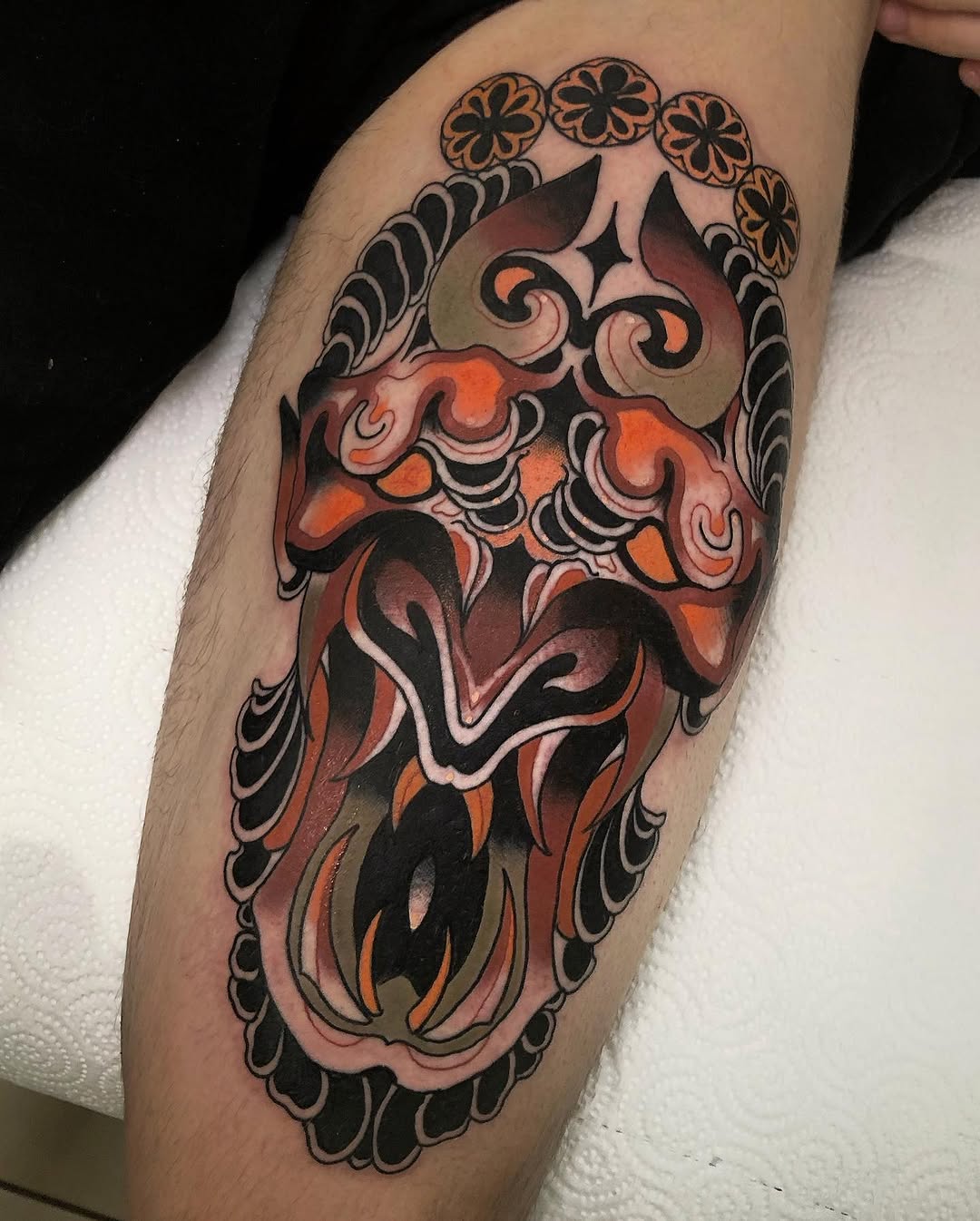 Colorful demon or oni mask tattoo on the calf featuring orange, red, and black tones with ornamental floral border details.