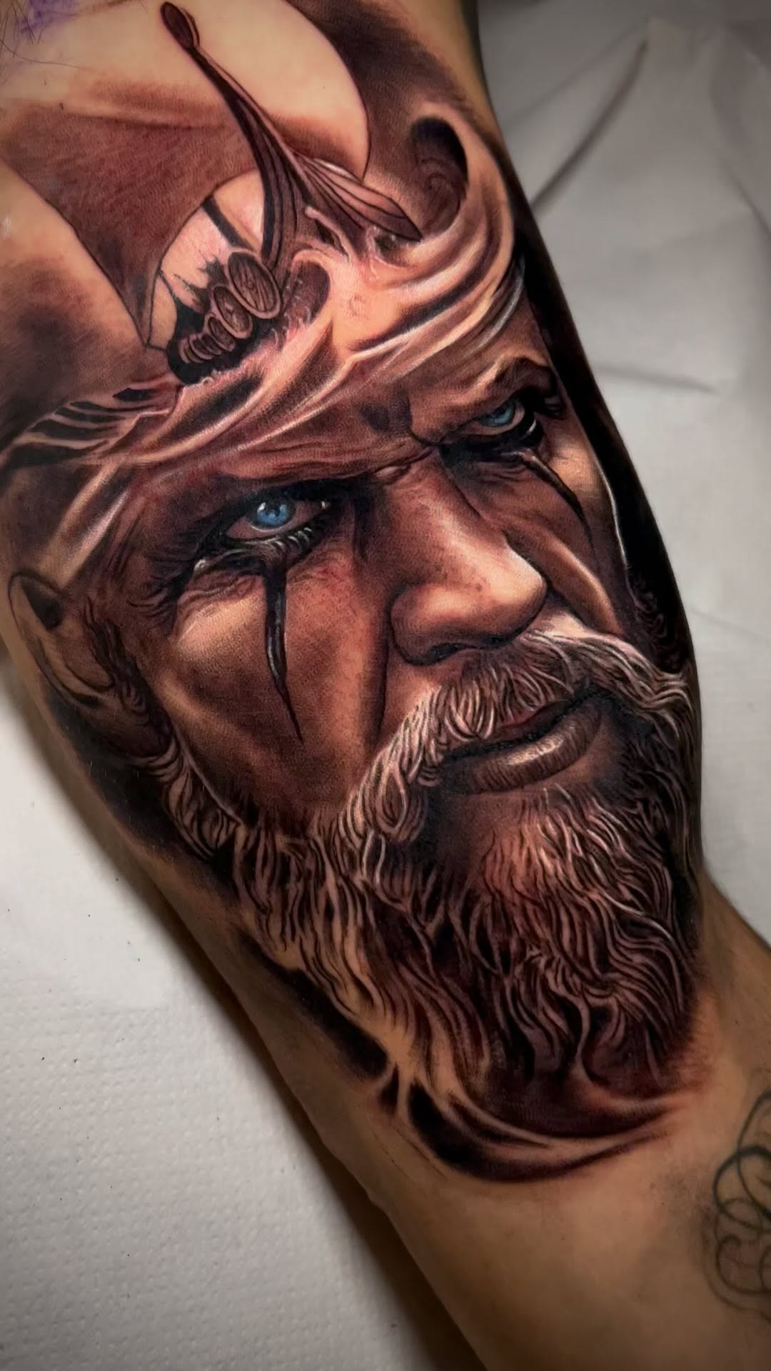 Detailed portrait tattoo of a bearded man's face with blue eyes and a small flying insect on his forehead, rendered in brown and tan tones on the arm.