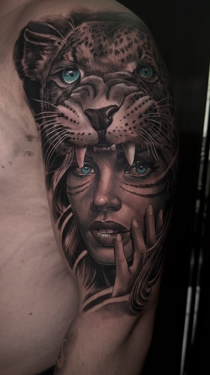 Shoulder tattoo depicting a woman's face with feline features and striking turquoise eyes, rendered in grayscale with detailed shading and linework.