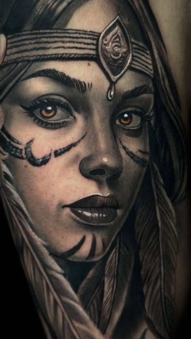 Detailed shoulder tattoo portrait of a woman with ornamental forehead jewelry, gold eyes, and decorative face markings in black and grey ink.