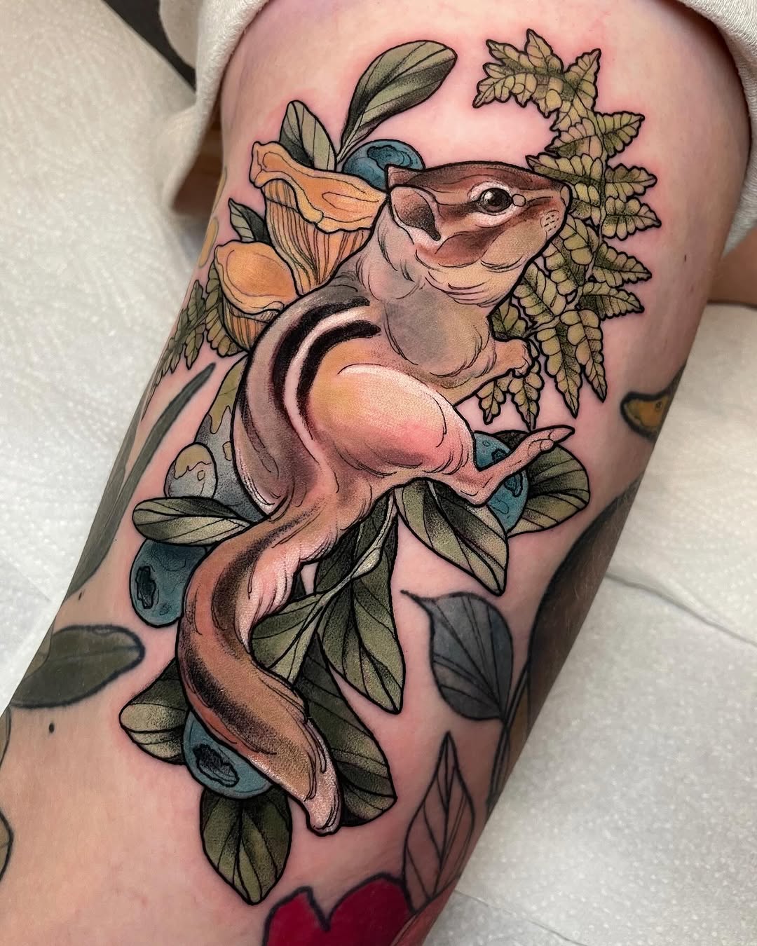 Colorful tattoo on upper leg featuring a smiling possum surrounded by flowering vines, leaves, and botanical elements in orange, green, and blue tones.