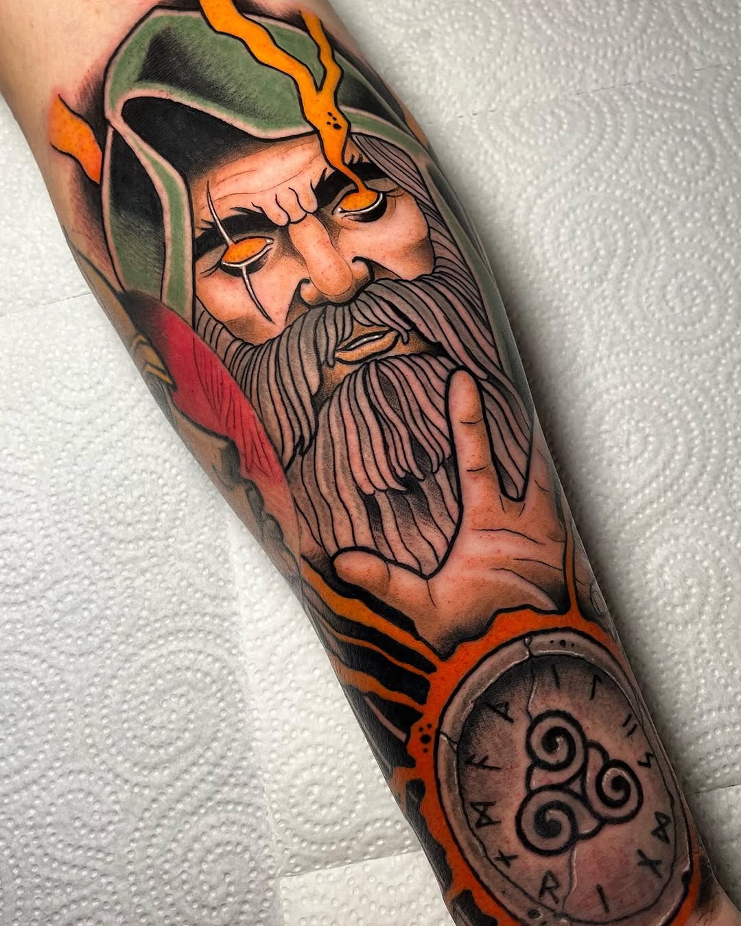 Colored arm tattoo featuring a bearded wizard with green hat and orange flames, paired with a spiral-marked pocket watch below.