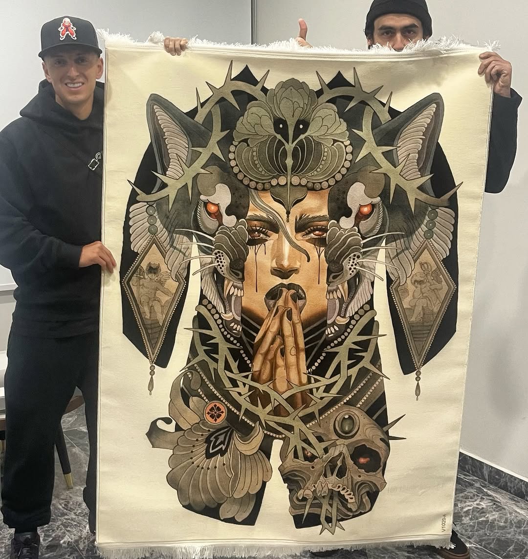 Two people hold a large fabric artwork featuring a symmetrical face with ornate wings, butterflies, skull, and botanical elements in sepia and black tones.