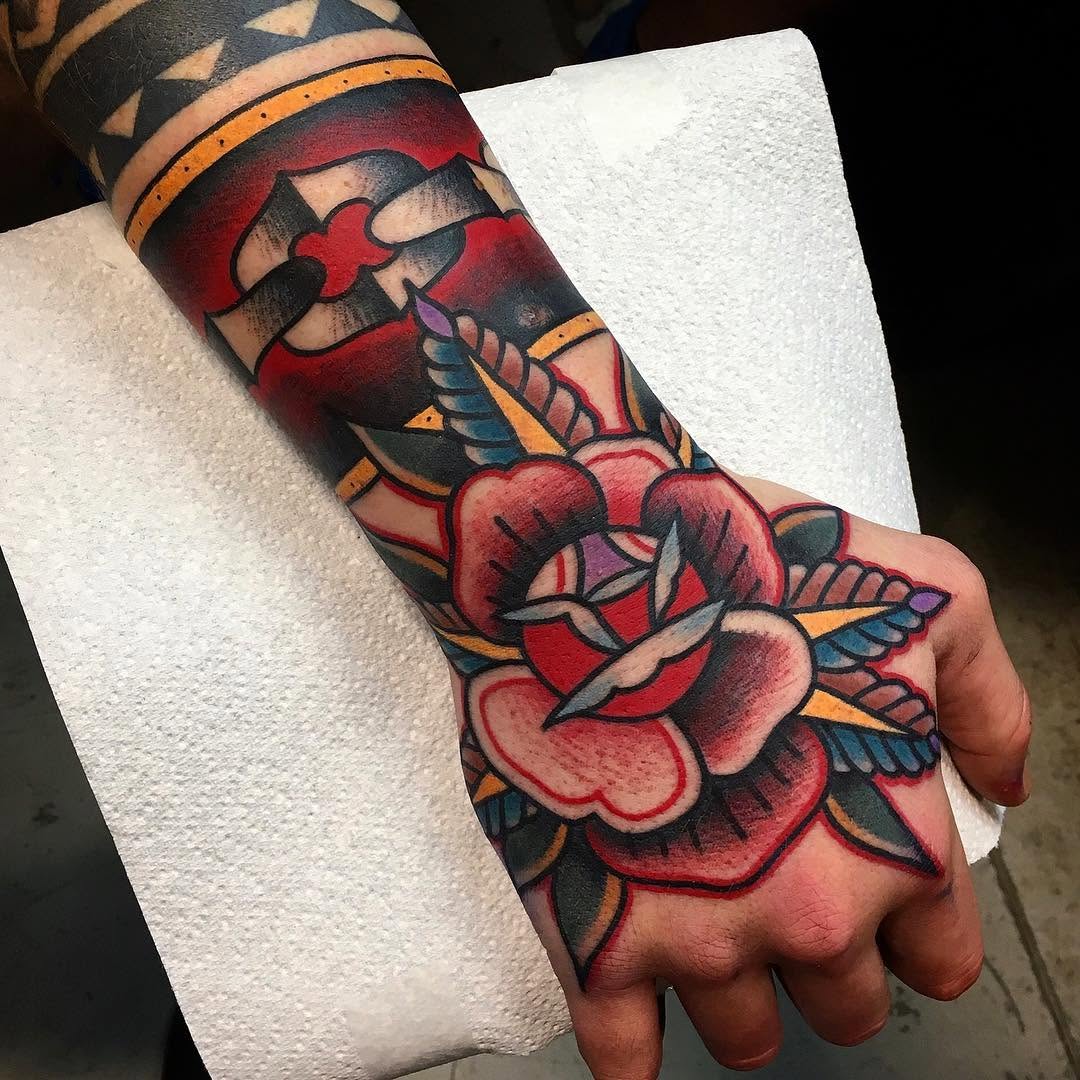 Colorful winged hearts and cross tattoos in red, black, orange, and blue covering the hand and forearm of a person holding white paper.