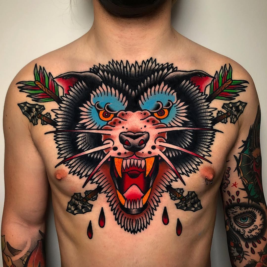 Colorful cat face tattoo on chest featuring blue eyes, orange markings, red mouth, black whiskers, and symmetrical floral and leaf elements on shoulders.