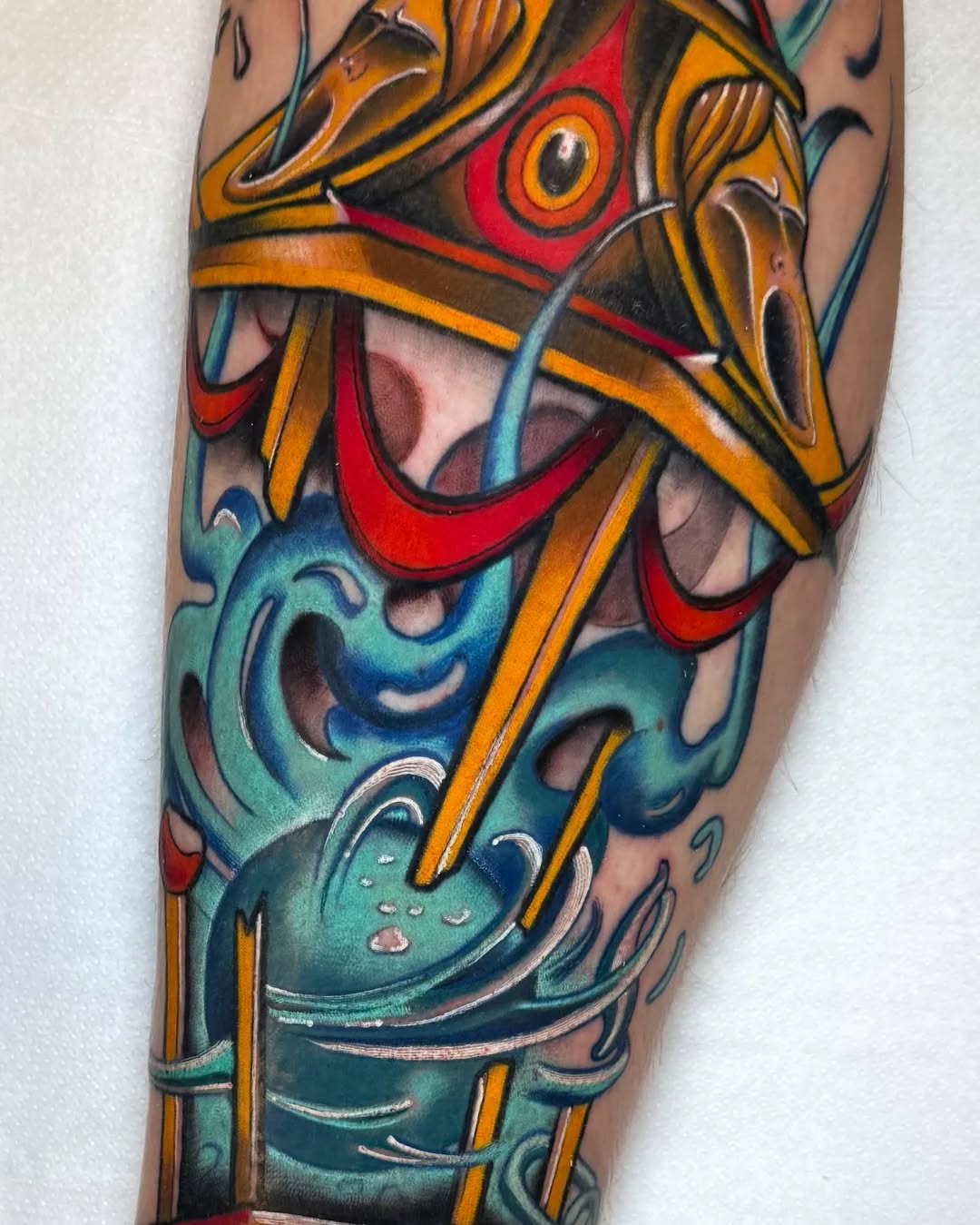 Colorful leg tattoo featuring stacked ornamental masks in red, gold, teal, and black with decorative swirling elements and striped accents.
