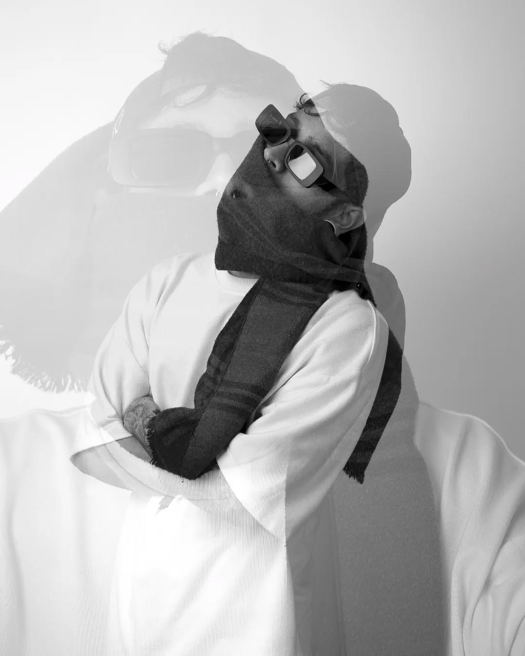 Person wearing white and black clothing with sunglasses displays a visible tattoo on their arm and neck area in a studio portrait.