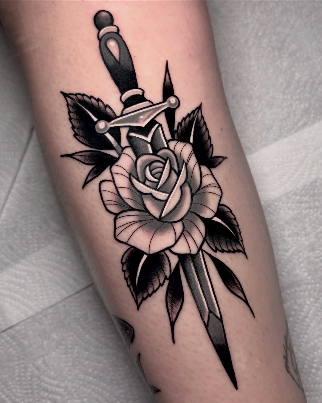 Dagger piercing through a rose with leaves tattooed on an arm in black ink with pink shading.