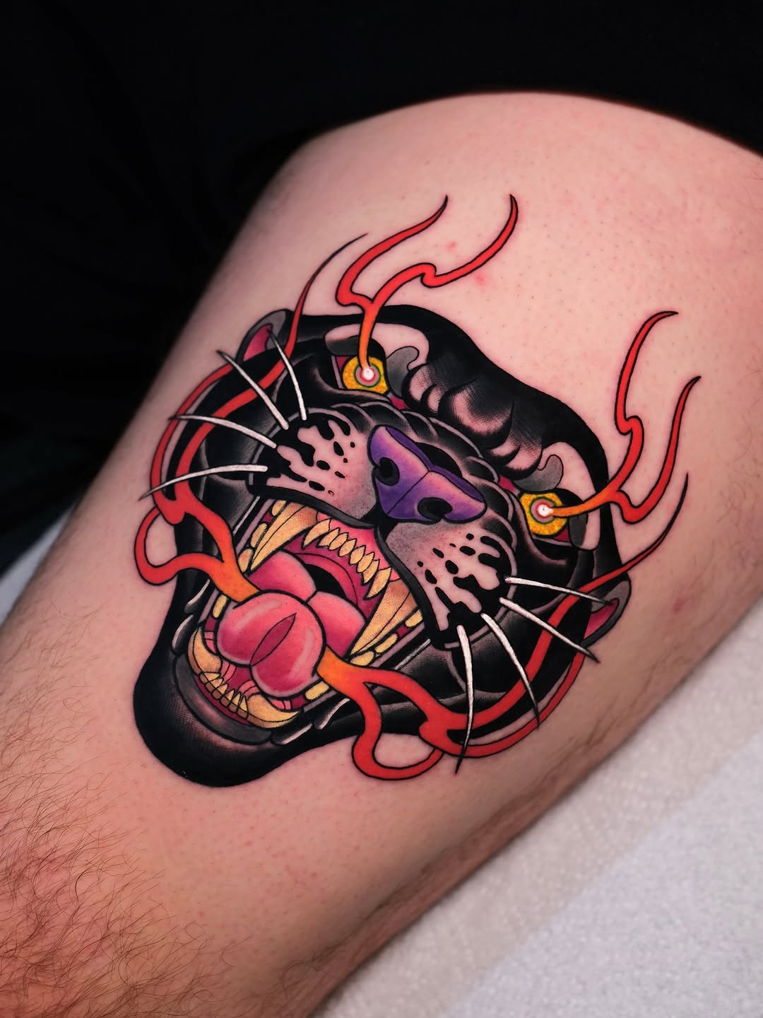 Colorful demon mask tattoo on upper arm featuring red flames, purple eyes, gold accents, and sharp teeth with pink mouth detail.