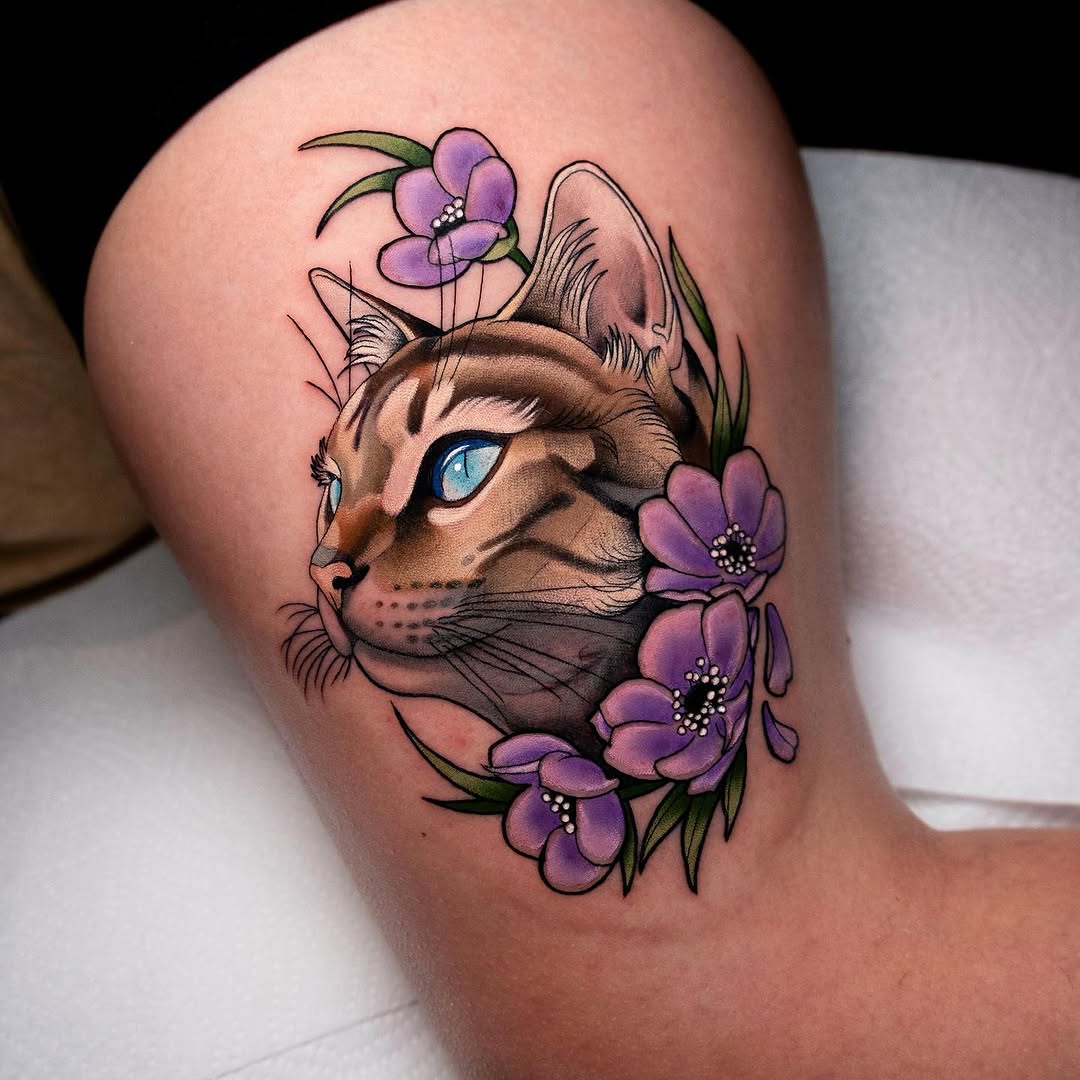A brown cat's head with blue eyes surrounded by purple flowers and green leaves, tattooed on the thigh.