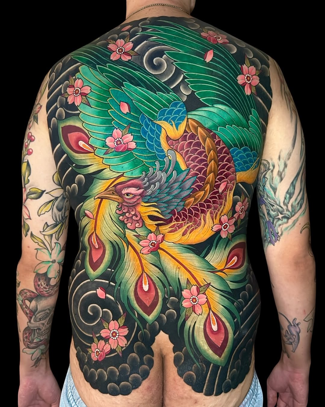 Colorful phoenix with spread wings and flowing tail feathers surrounded by pink flowers and decorative clouds covers the entire back of a shirtless man.