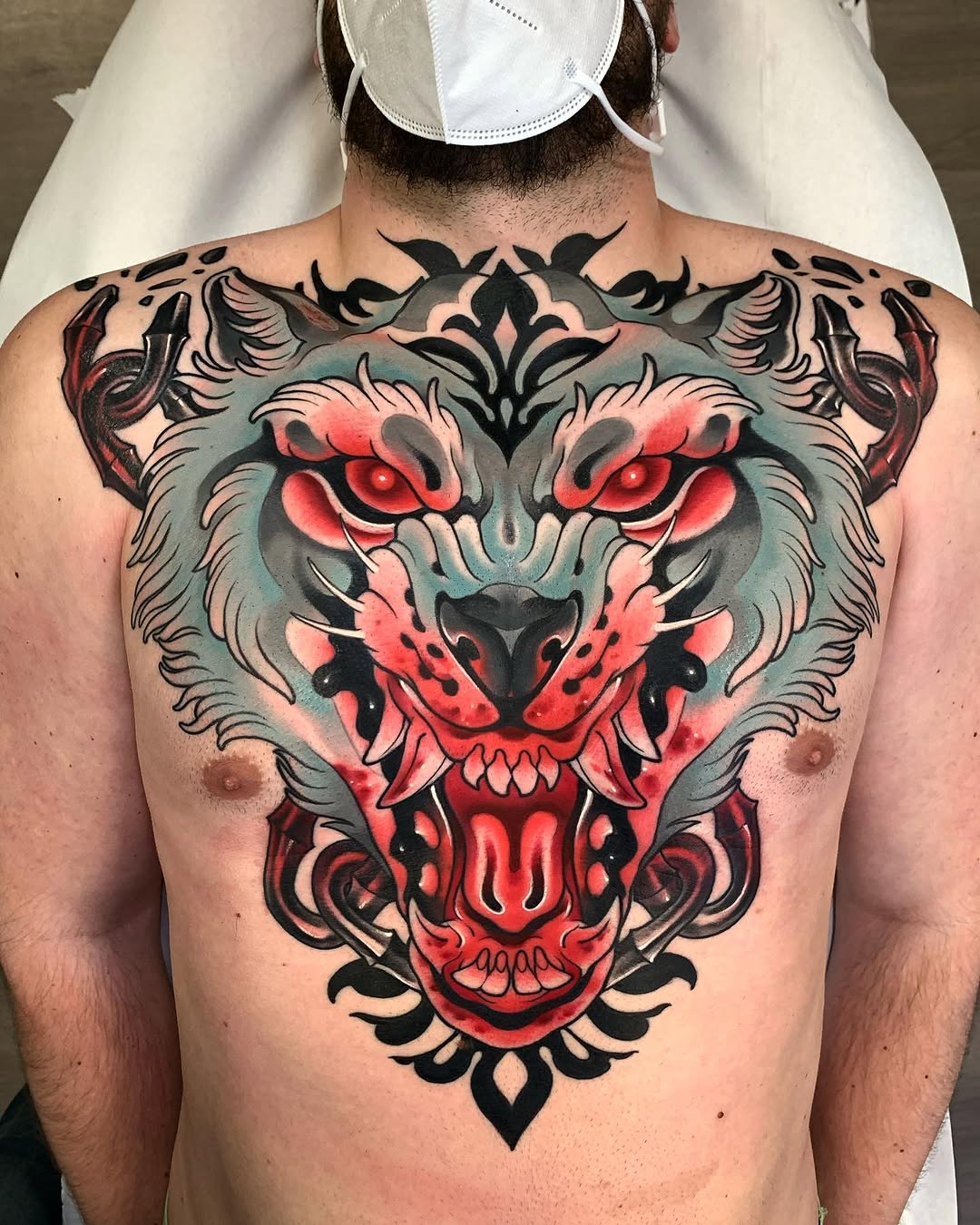 Large symmetrical tiger face and skull design on chest in red, black, and teal ink with ornamental flourishes extending across shoulders.