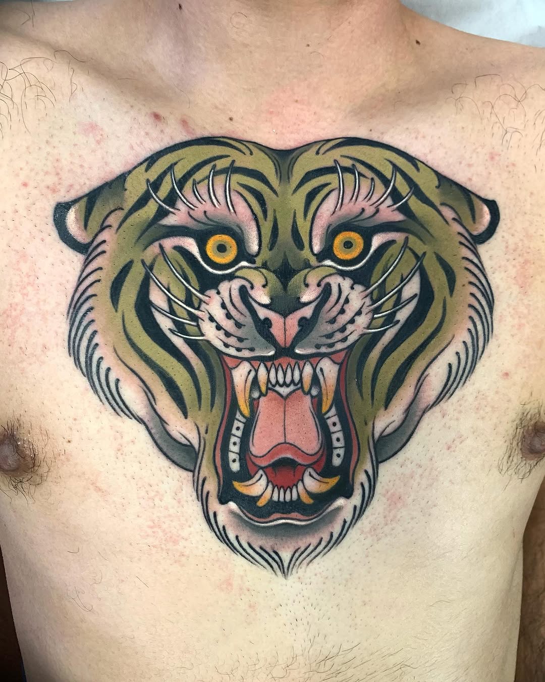 Tiger head chest tattoo featuring golden and olive green stripes, bright yellow eyes, pink nose, open mouth with visible fangs and teeth, and red inner mouth details.