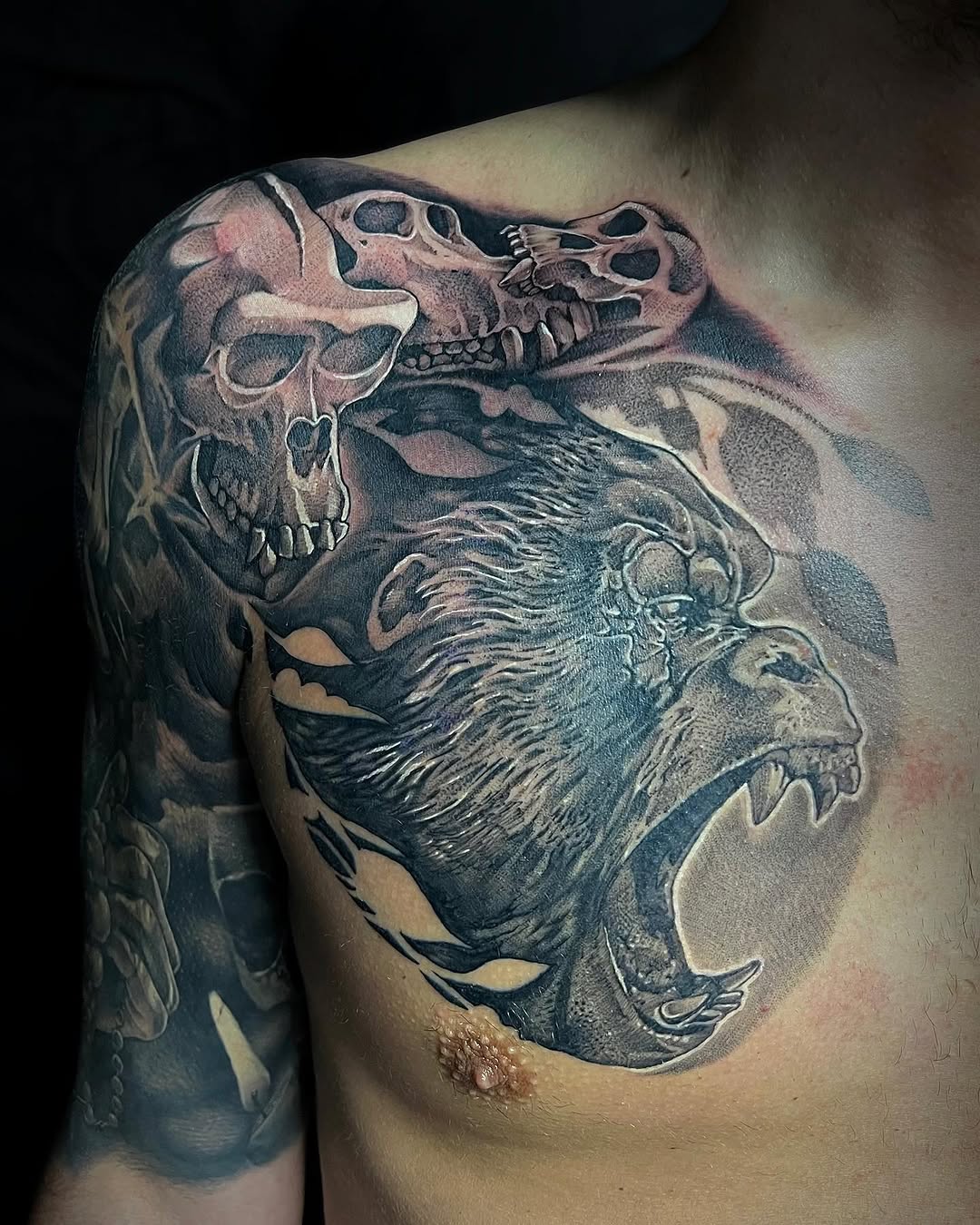 Shoulder tattoo depicting three skulls and a bearded male profile with flowing hair, rendered in dark grey and black ink with shading.