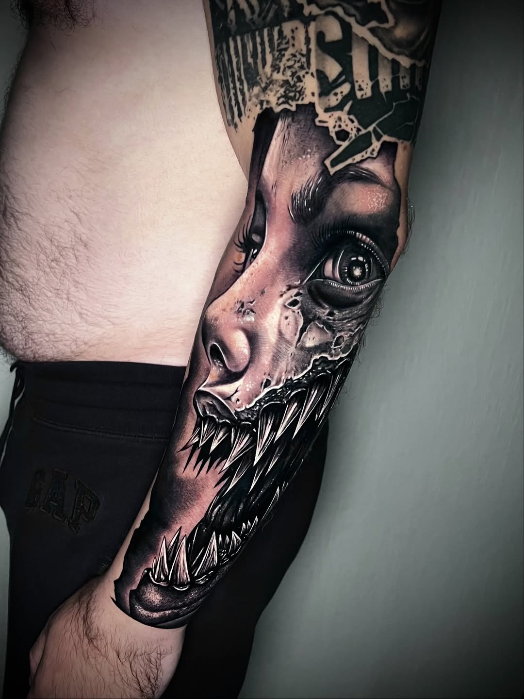Detailed demon face tattoo on upper arm with sharp teeth, piercing eye, and monochromatic shading in black and gray tones.