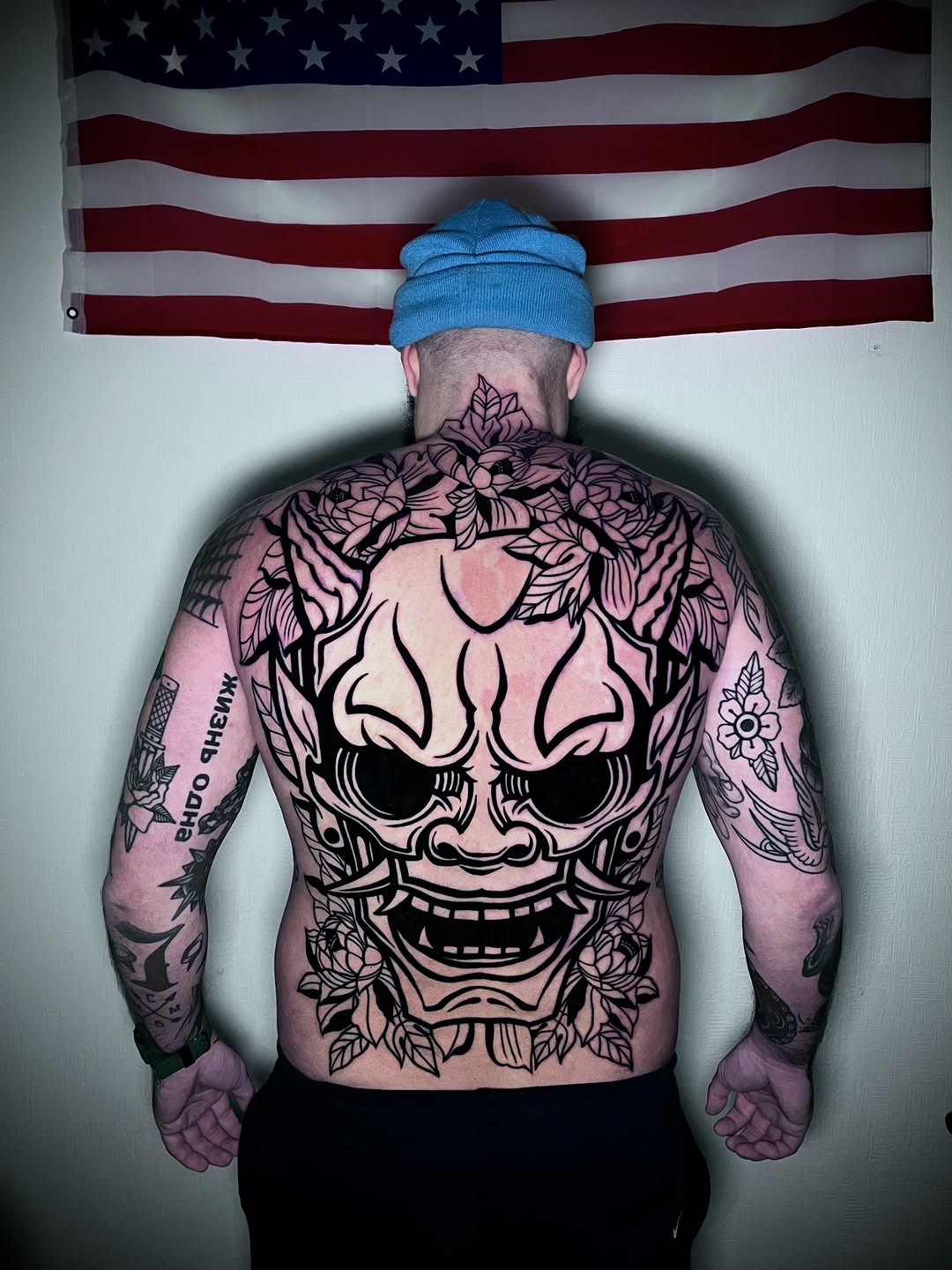 Shirtless man with large skull and floral design covering entire back, surrounded by additional tattoos on arms and neck, posed against American flag.