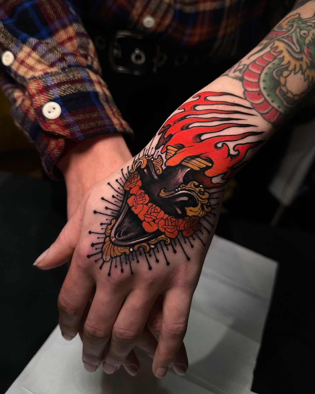 Colorful hand tattoo featuring a black bird with red roses, flames, and ornamental details, extending up the forearm.