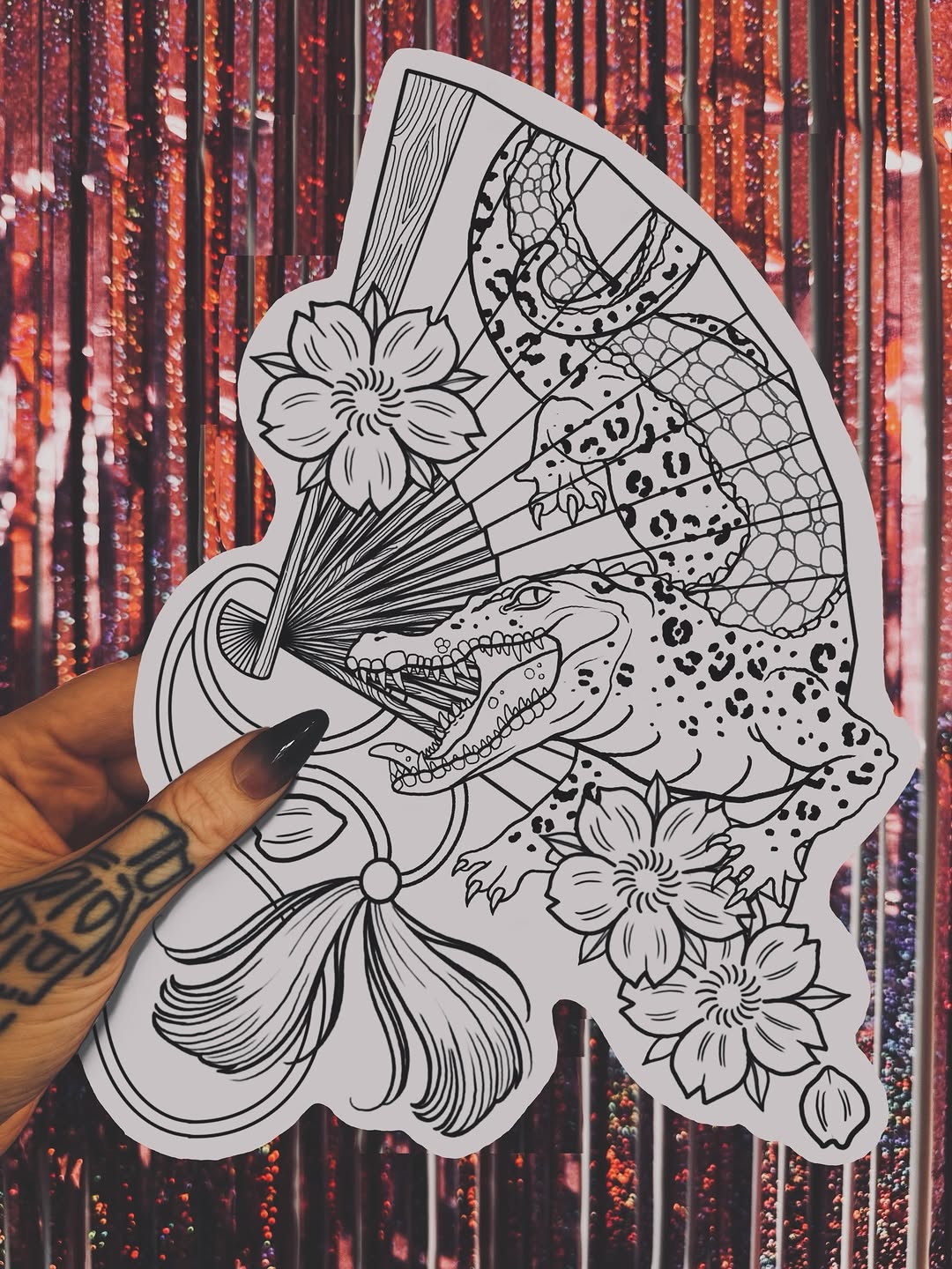Detailed line drawing of a crocodile with floral elements and patterned wings held by a tattooed hand against a red sequined backdrop.