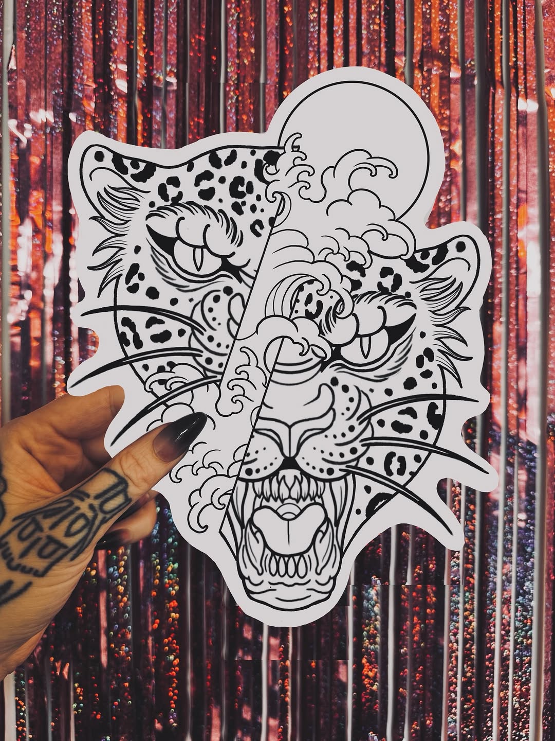 Hand holding an outlined sticker design featuring a layered leopard, cloud, and tiger face composition with intricate line work and dot patterns.