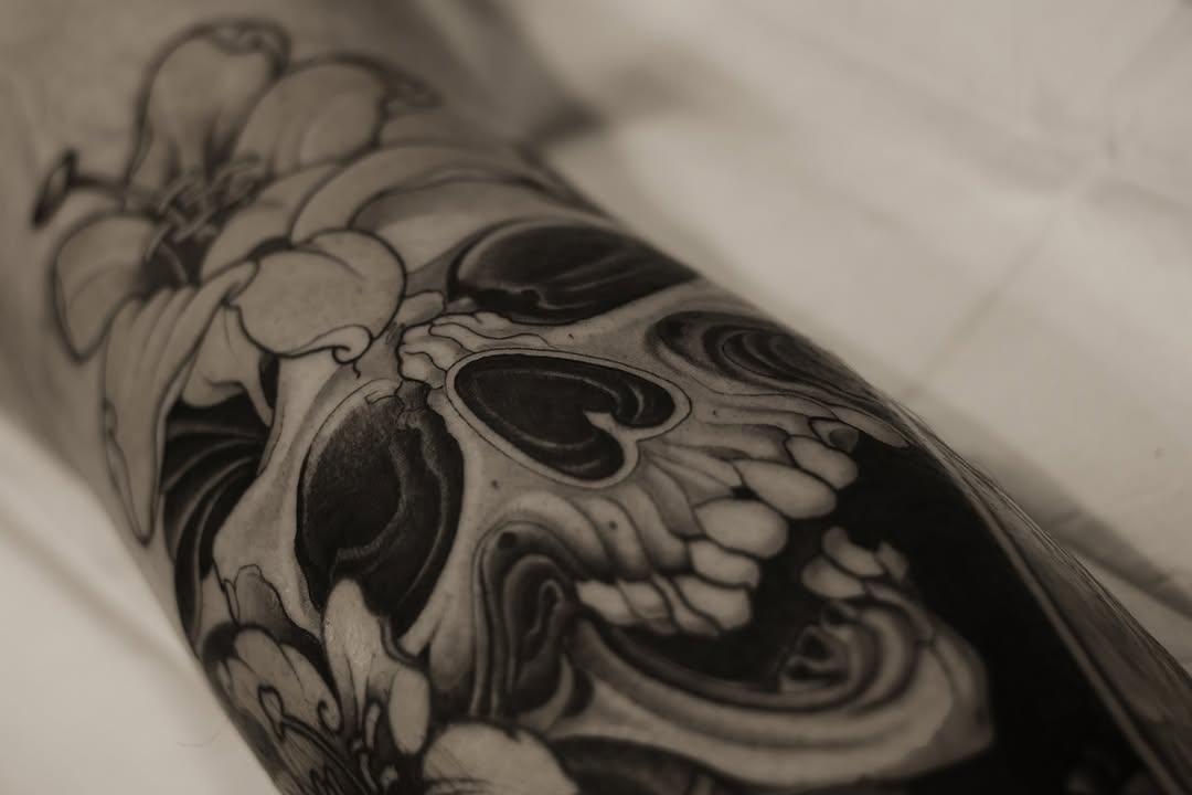 Detailed skull tattoo with floral elements and intricate shading on the forearm, rendered in black ink with varying gray tones.