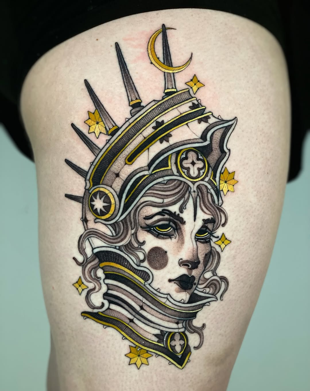 Ornate feminine face with wavy hair, crescent moon and spear crown, stars, and cross details in black, gold, and gray on upper thigh.