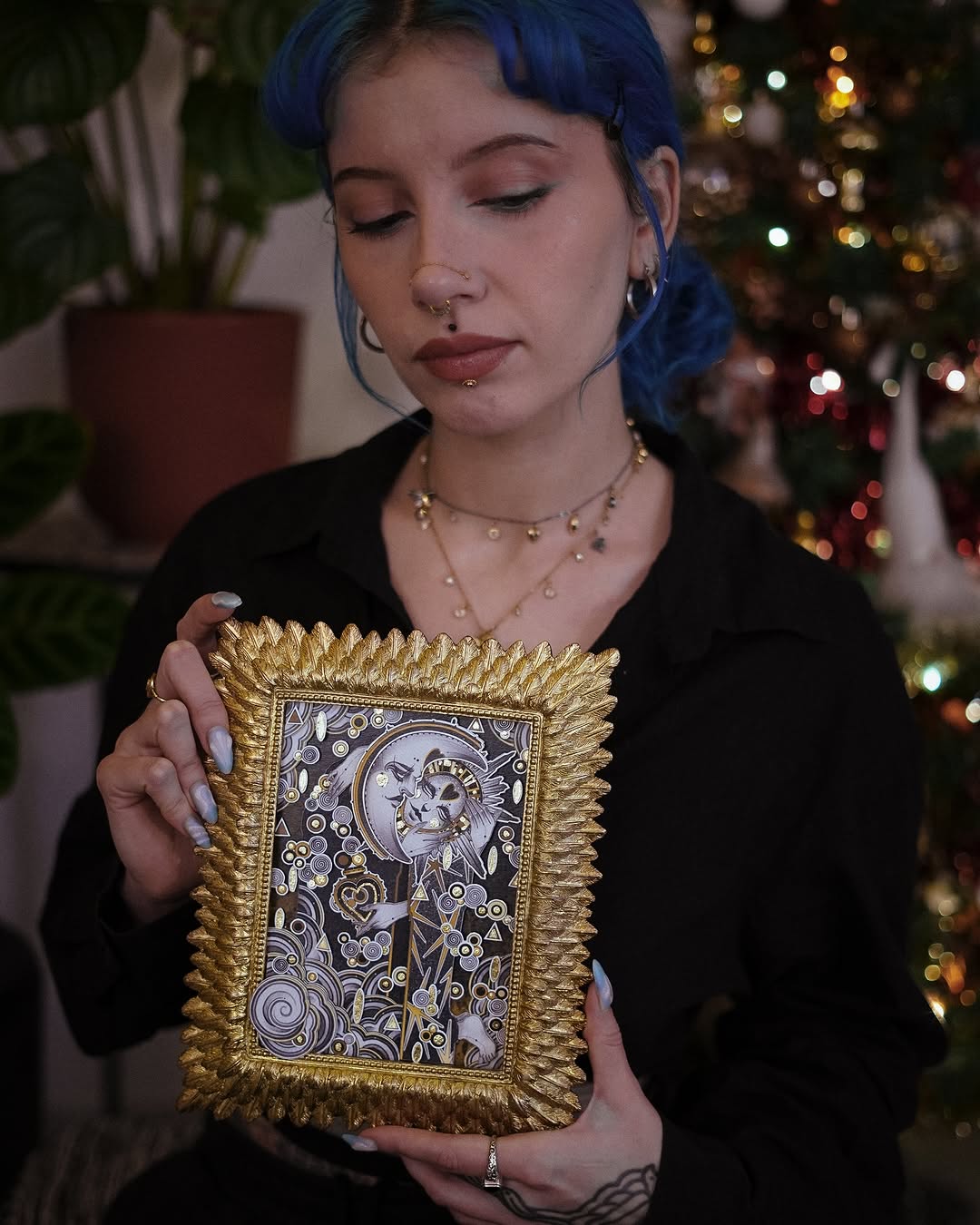 Woman with blue hair displaying a framed arm tattoo featuring a moon-faced figure surrounded by decorative swirls and ornamental elements in black and gold tones.
