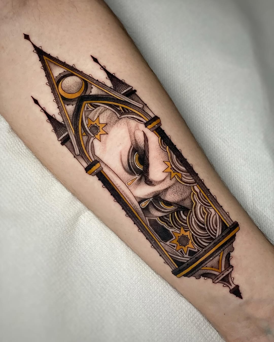 Celestial face tattoo in geometric frame with moon, stars, and flowing hair details in black, gold, and tan on forearm.