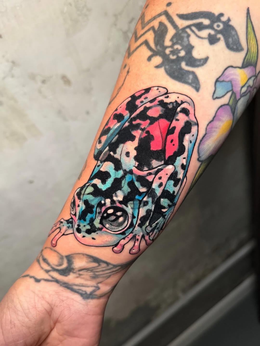 Colorful spotted frog or toad tattoo on forearm with pink, blue, and black patches, surrounded by smaller black silhouette designs on the arm.