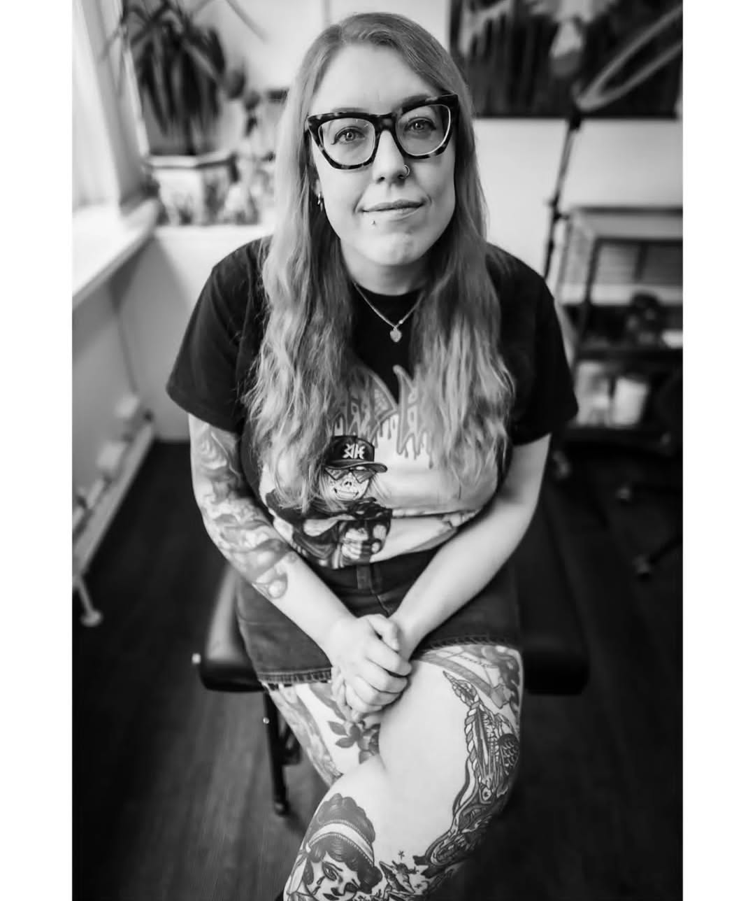 Portrait of a tattooed woman with glasses wearing a graphic t-shirt, displaying arm and leg tattoos in black ink in an indoor studio setting.