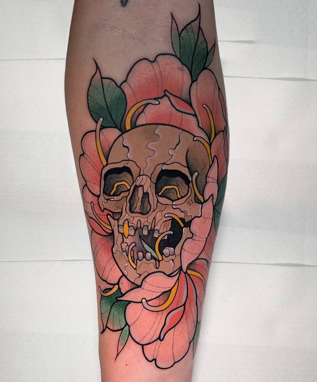 Skull with open mouth surrounded by large pink flowers with green leaves and gold accents, tattooed on the calf.