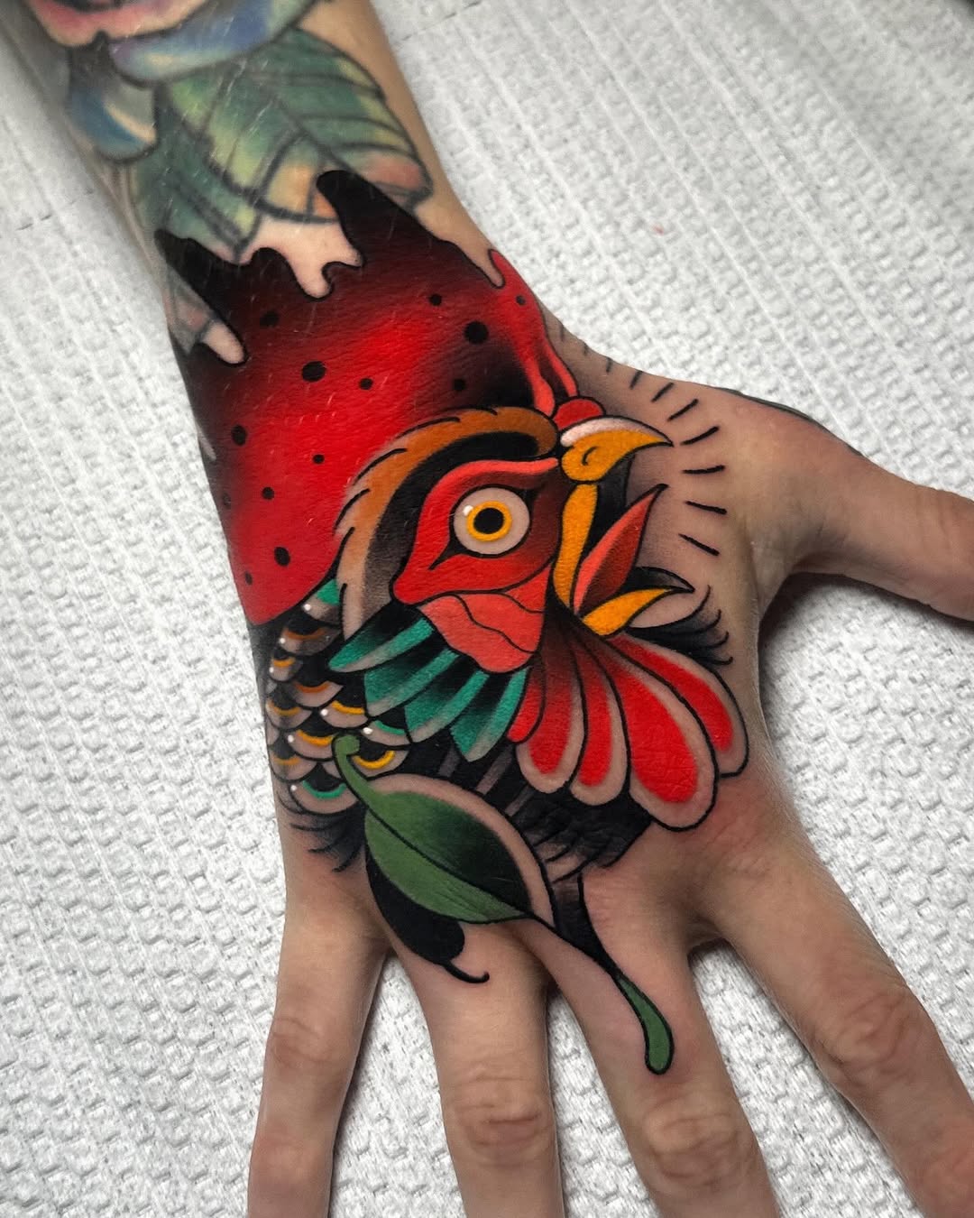 Colorful bird tattoo on the back of a hand featuring red plumage, orange beak, green and yellow wing details, and a leafy stem below.
