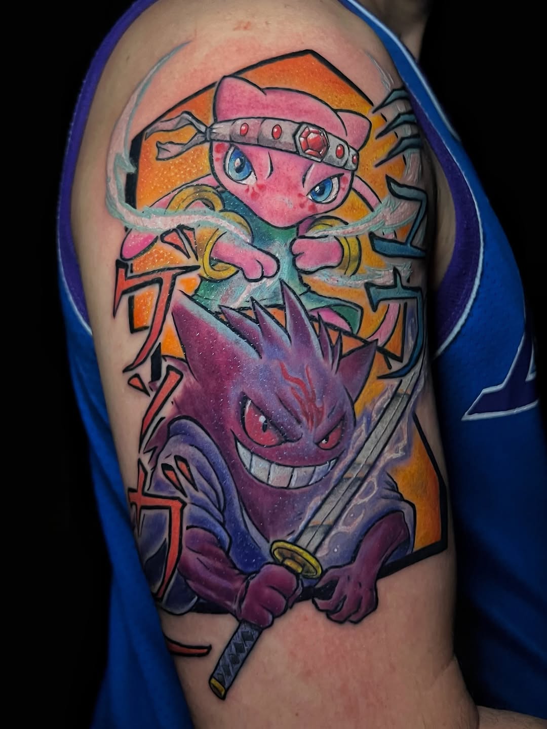 Colorful shoulder tattoo featuring a pink character with a headband and a purple demon face below, holding a sword, with orange and yellow background elements.
