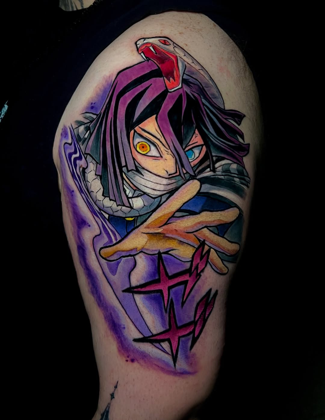 Colorful shoulder tattoo featuring an anime character with mismatched eyes, bandaged face, purple and magenta tones, and crossed arms with decorative symbols.