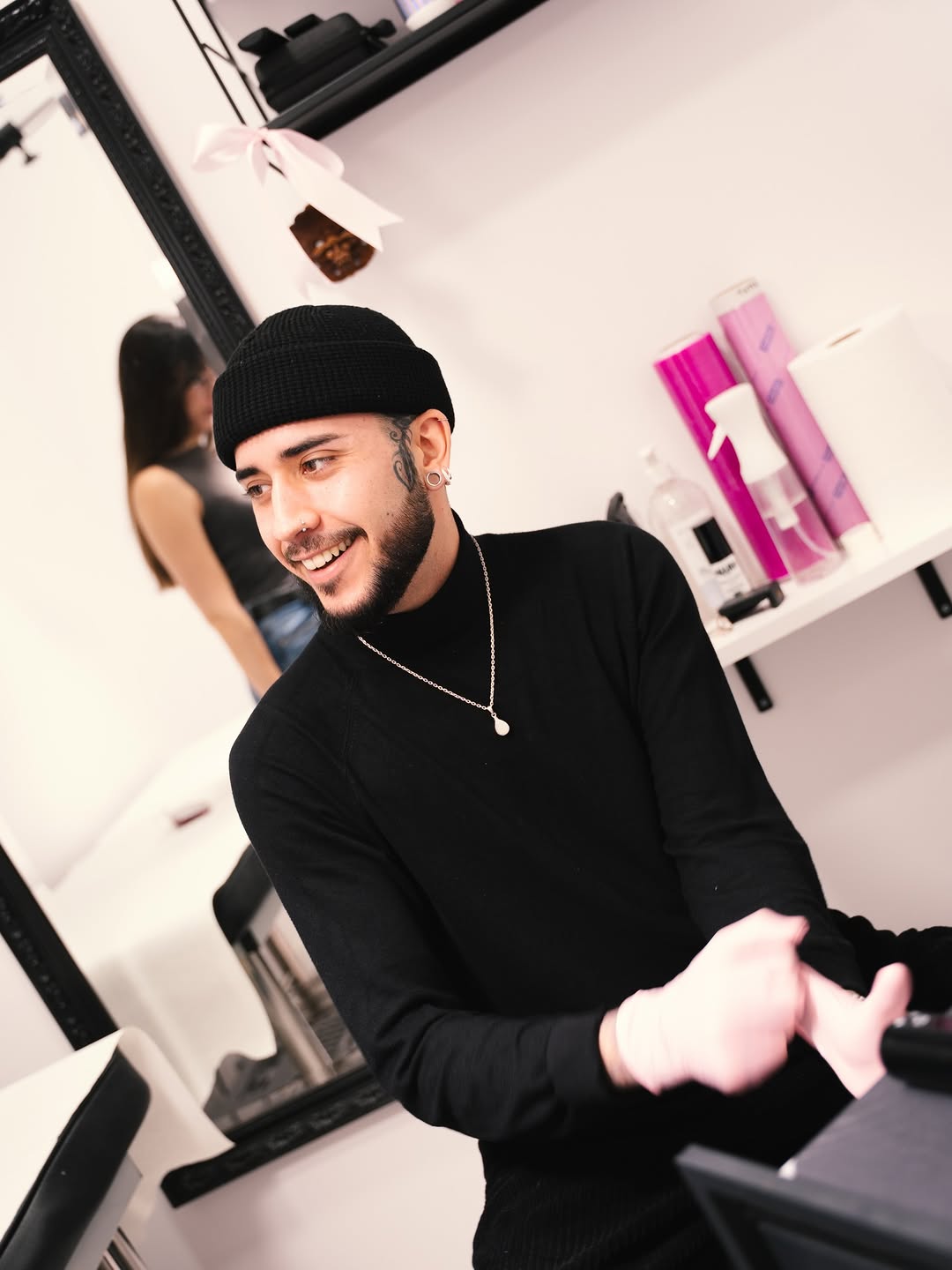 Smiling man with a small ear piercing wearing a black beanie and necklace in a tattoo studio with pink product bottles visible on the shelf behind him.