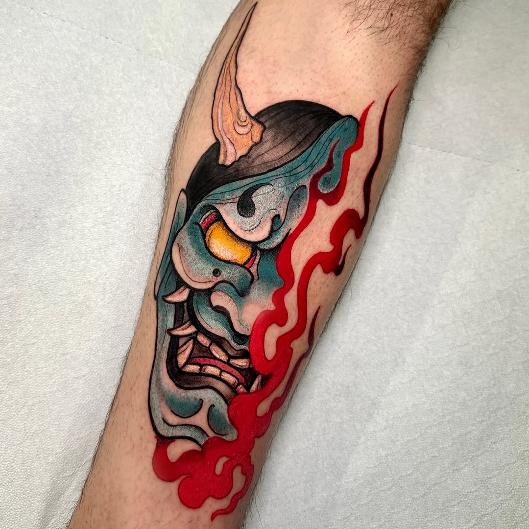 Colorful demon or oni mask head tattoo on a calf with teal, red, yellow, and black colors, featuring flames trailing downward.