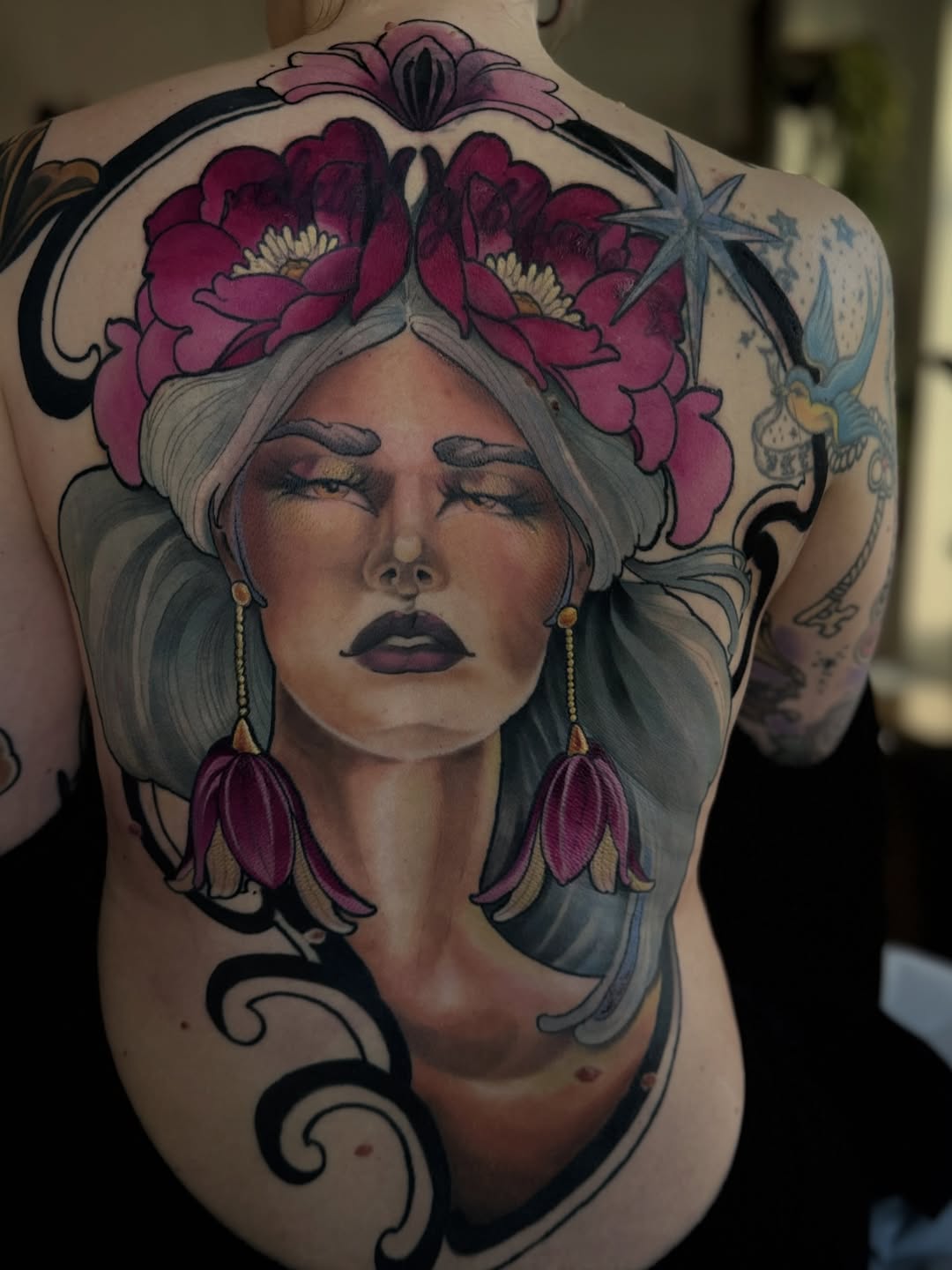 Large-scale back tattoo featuring a woman with gray hair, closed eyes, burgundy flowers in her hair and as dangling earrings, with decorative black swirls and blue stars.