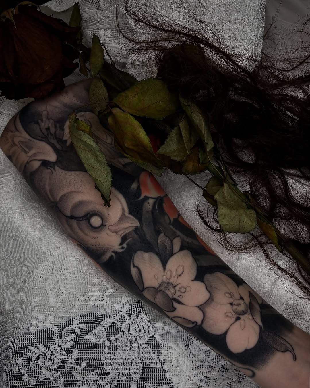 Arm tattoo featuring floral and botanical elements in black and gray ink, displayed on light fabric with dried flowers.