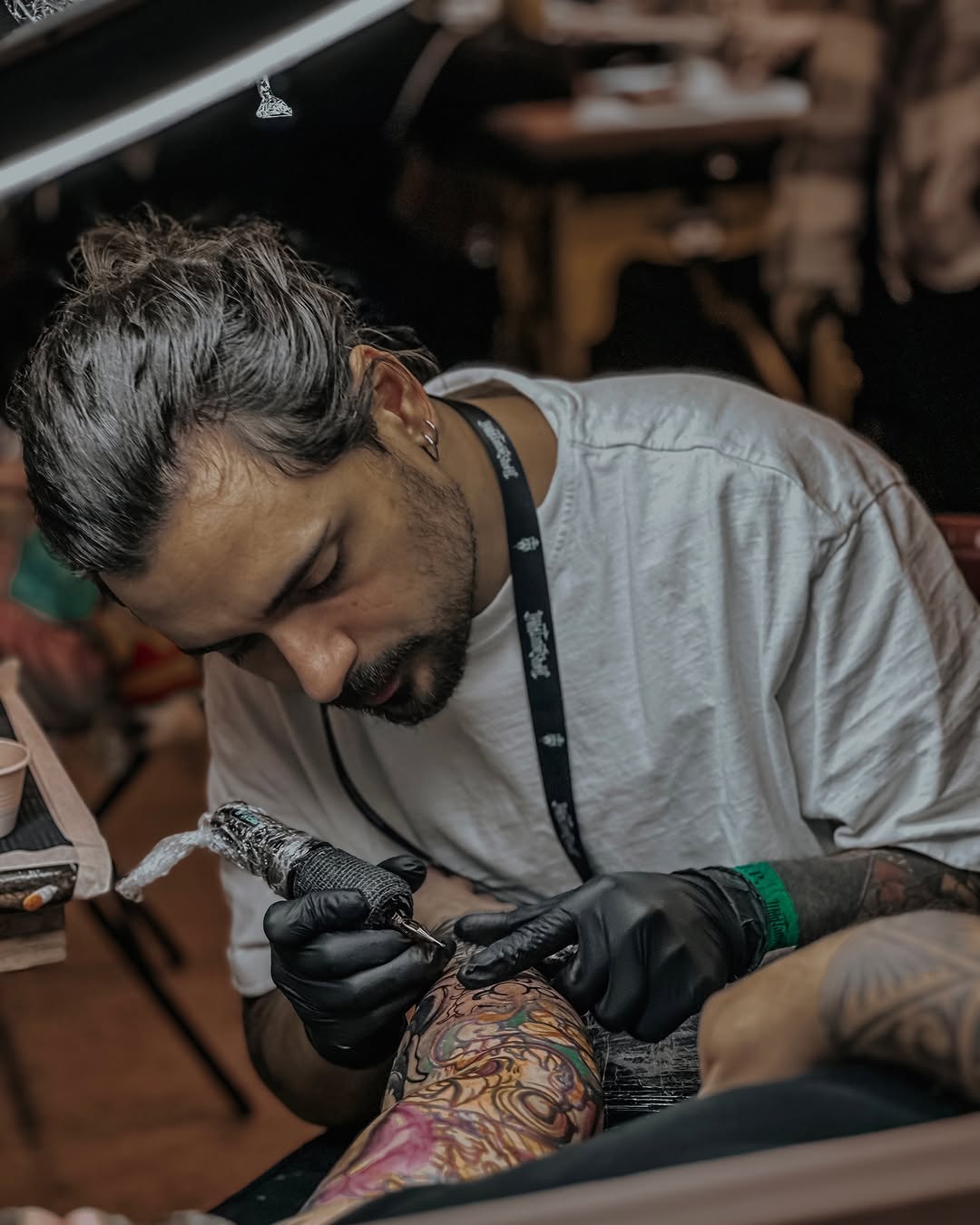 Tattoo artist wearing black gloves and white shirt applies colorful ink to a client's arm decorated with existing floral and abstract designs.