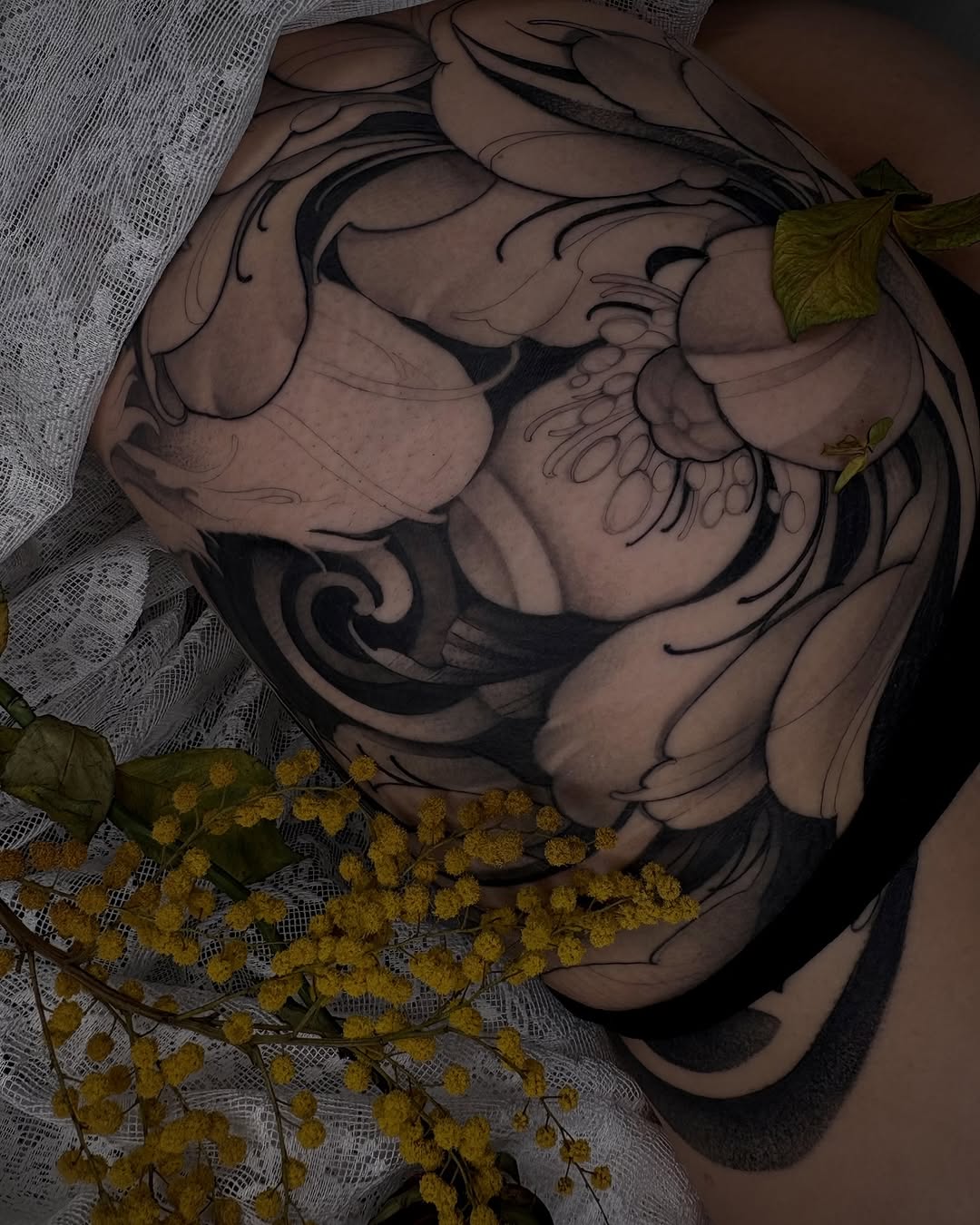 Detailed skull tattoo on chest with black linework and shading, displayed on skin with yellow dried flowers and white mesh fabric.