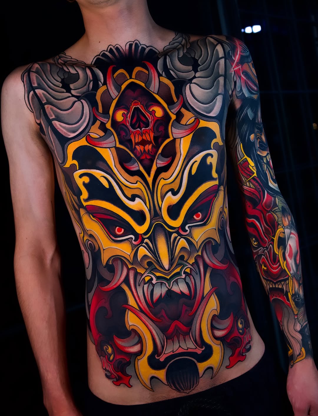 Colorful chest and sleeve tattoo featuring stacked demon faces with skull imagery, ornate gold scrollwork, and red accents on bare-chested male torso.