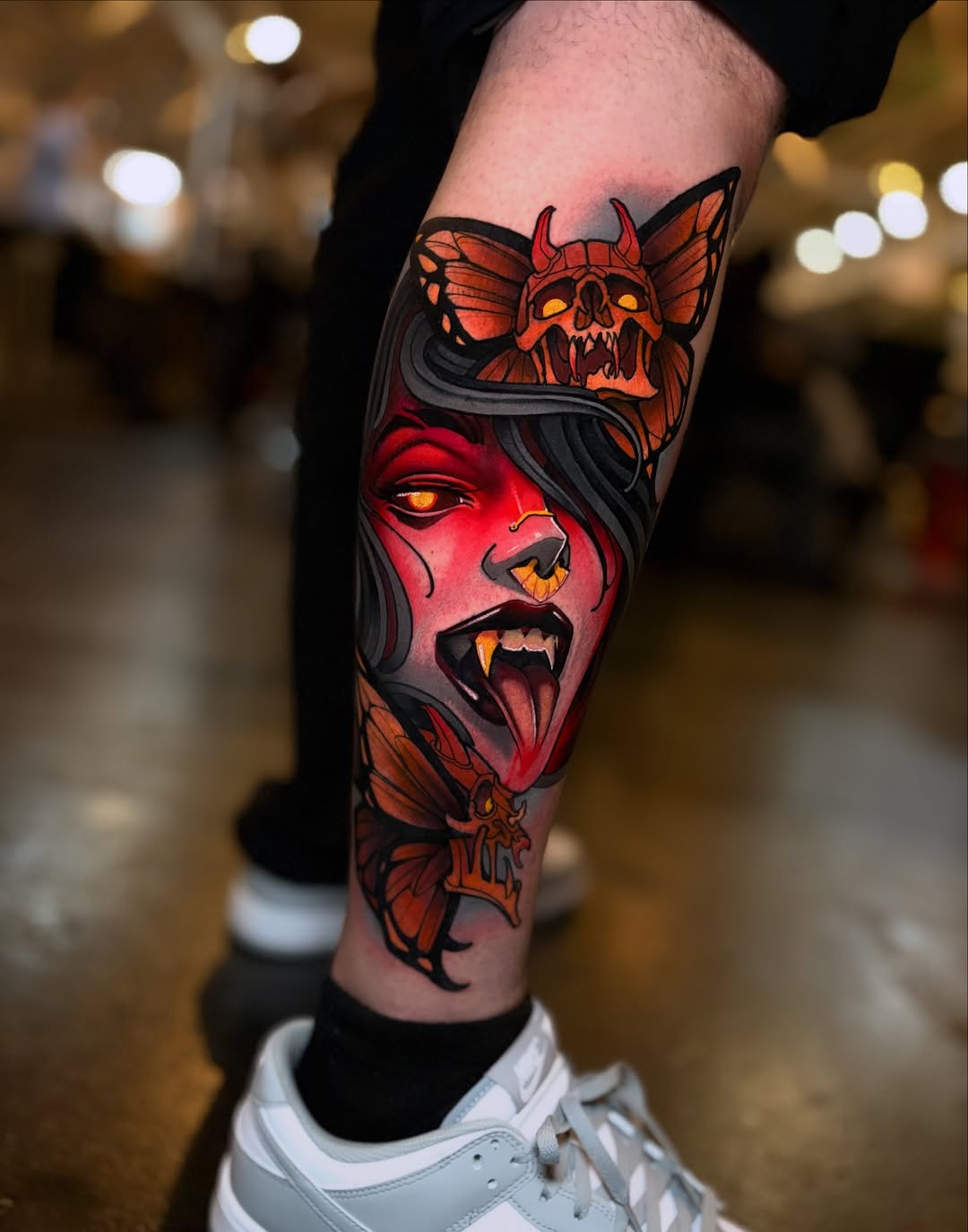 Red and orange demonic female face with horned skull above and butterfly motifs on the calf, featuring glowing yellow eyes and dark outlines.