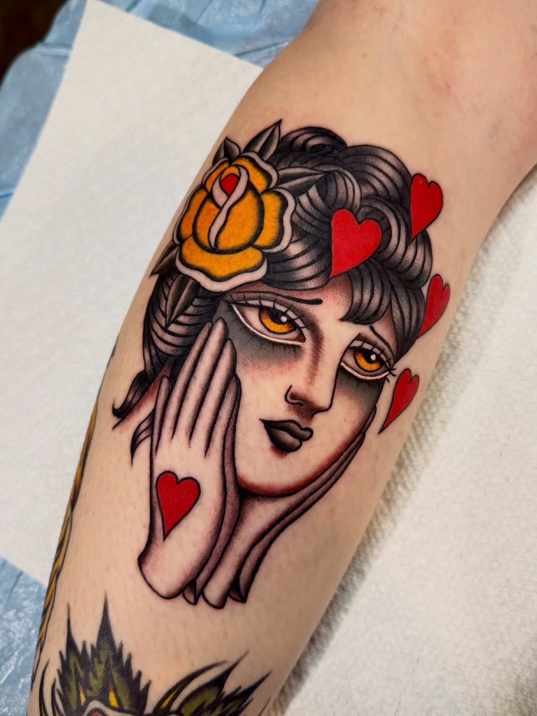 Female portrait tattoo on forearm featuring golden eyes, orange flower crown, red hearts, dark hair, and black outlines with color accents.