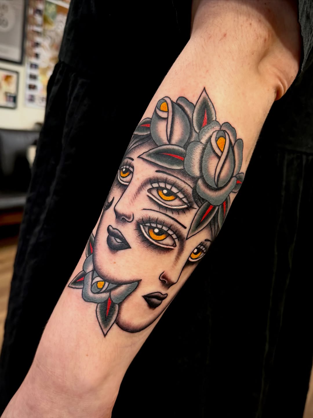 Portrait tattoo on forearm featuring a face with multiple eyes in golden yellow, gray flowers, and red accents throughout the design.