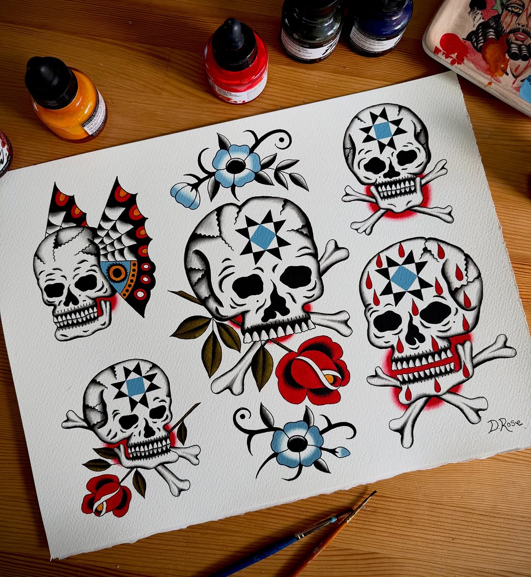 Tattoo flash sheet featuring five skulls with crossbones, geometric patterns, and red and blue flowers on white paper with watercolor paints nearby.