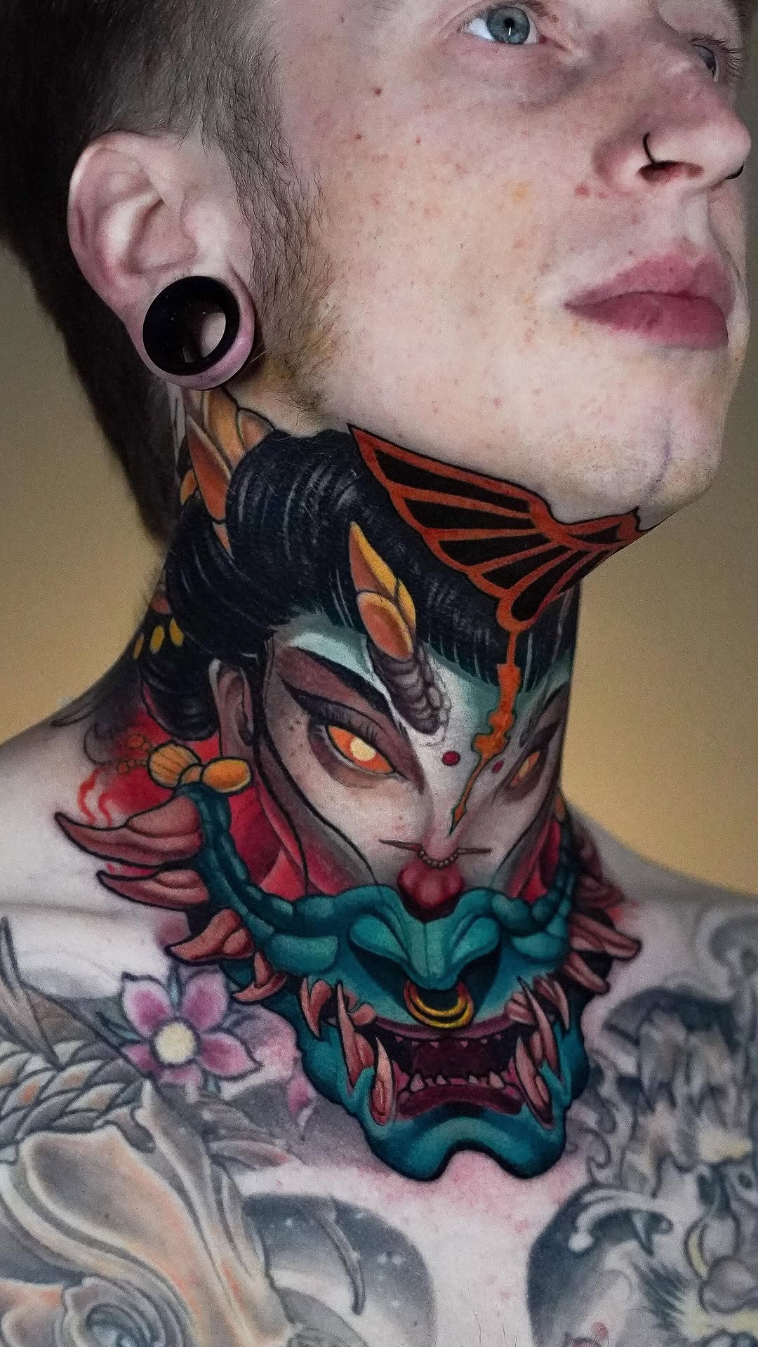 Colorful neck tattoo featuring a stylized demon face with red, teal, and orange details, flanked by floral and butterfly elements.