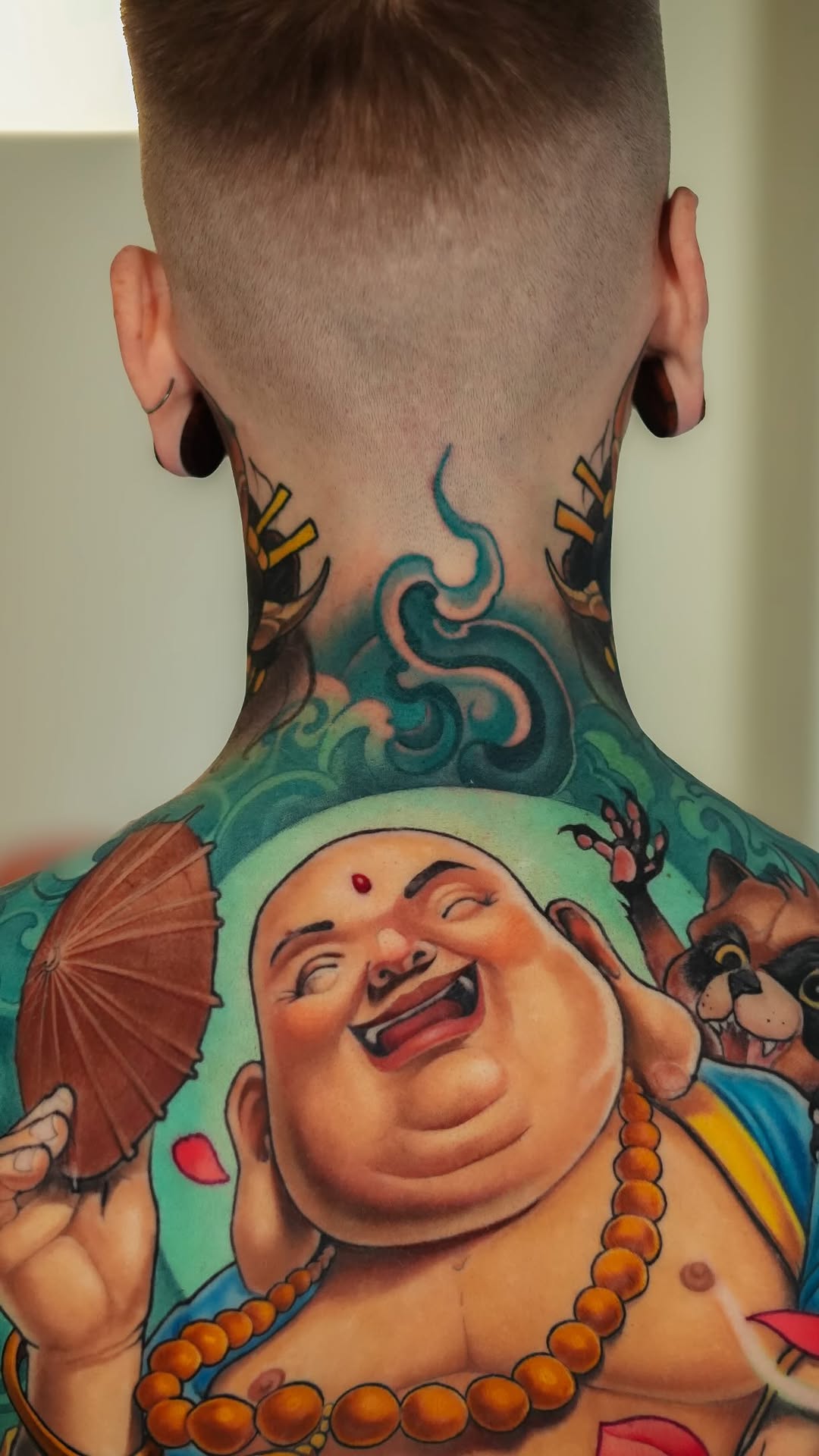 Back of neck and upper back tattoo featuring a laughing Buddha figure with gold beads and ornamental designs in teal, orange, and skin tones.