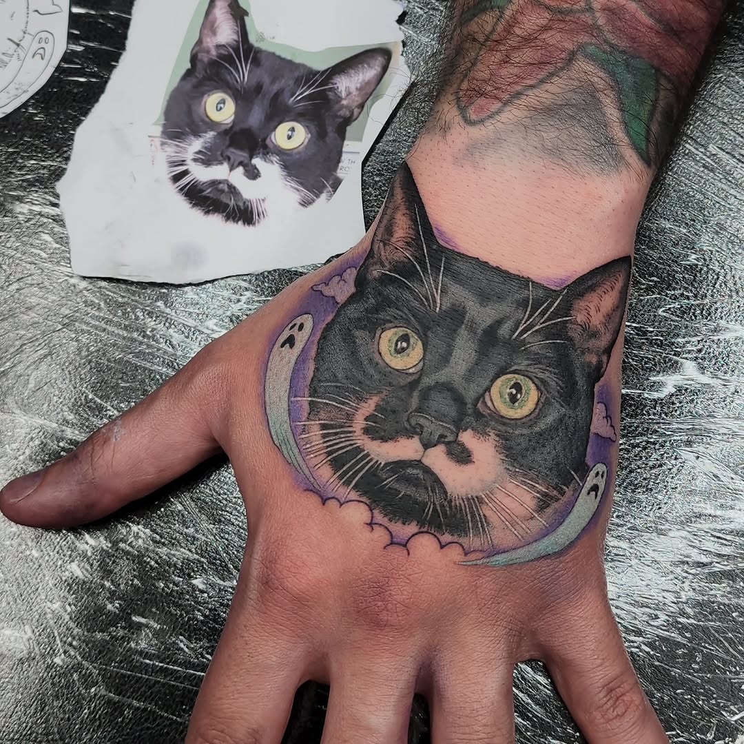 Black and white cat face tattoo on the back of a hand with purple and green crescent moon framing and small decorative details.