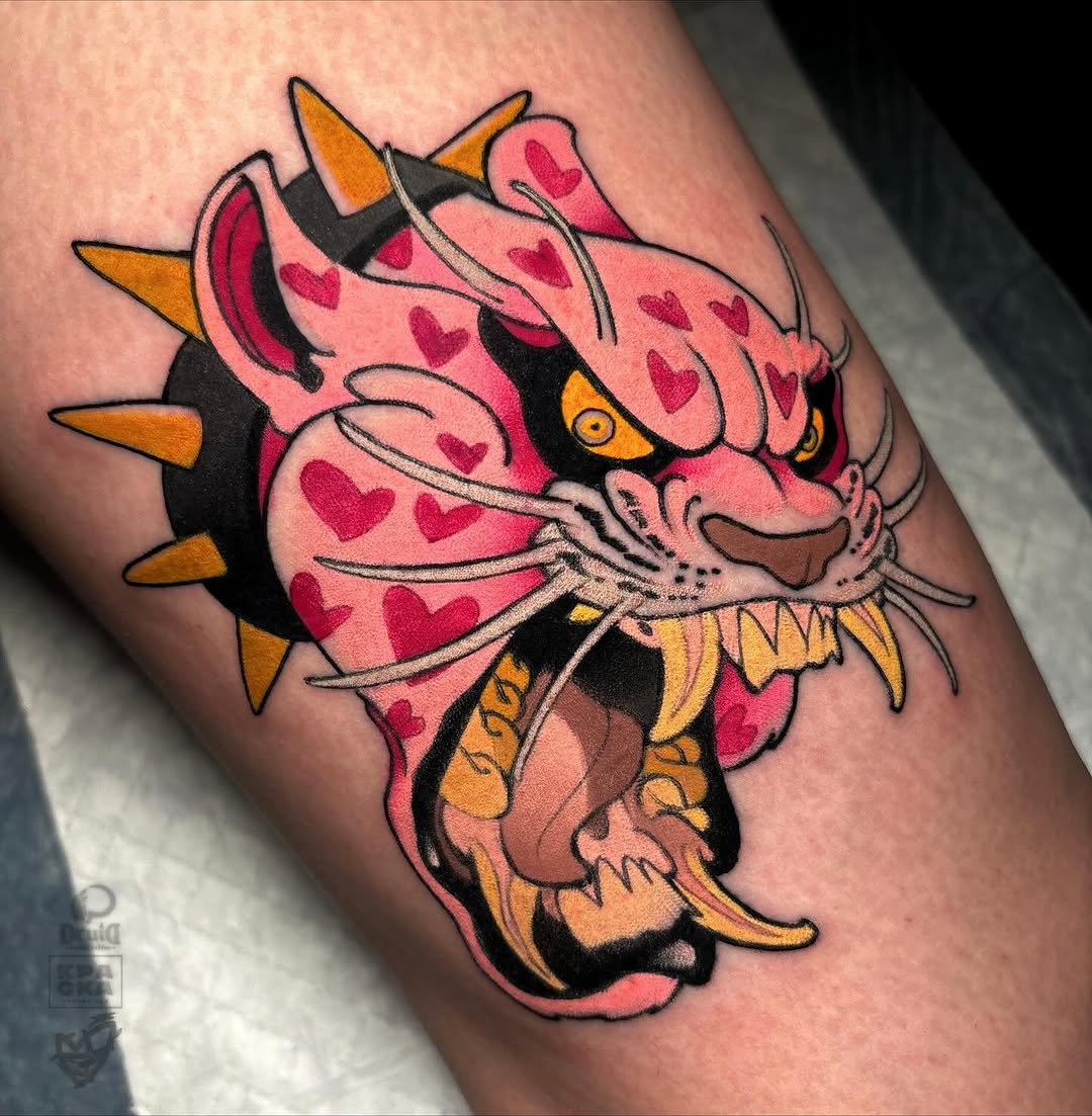 Pink demon or oni head with red hearts, gold spikes, and yellow eyes tattooed on an upper arm in red, pink, black, and gold colors.